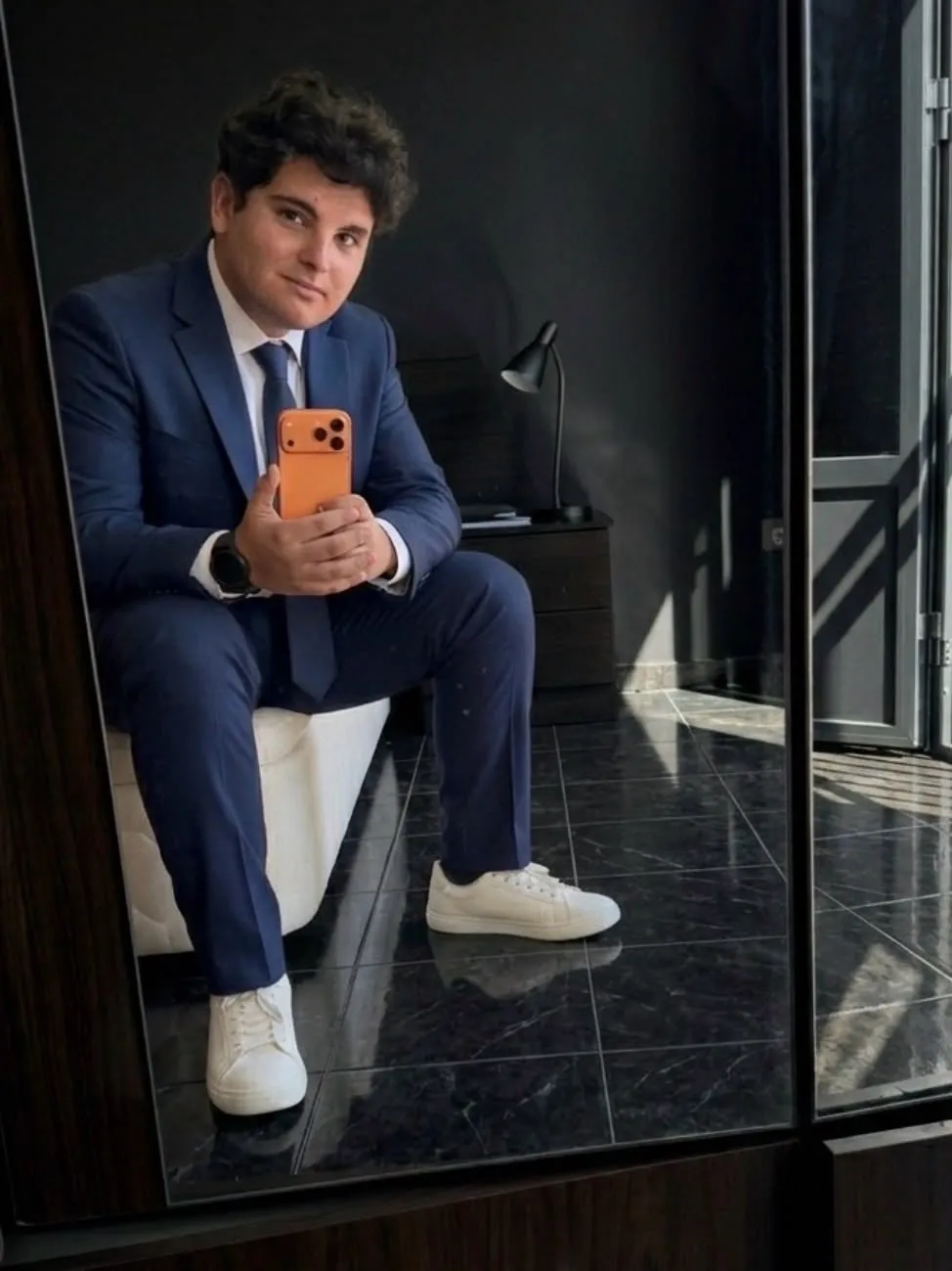 Aaron Alba con traje azul, sentado, haciéndose una foto frente a un espejo