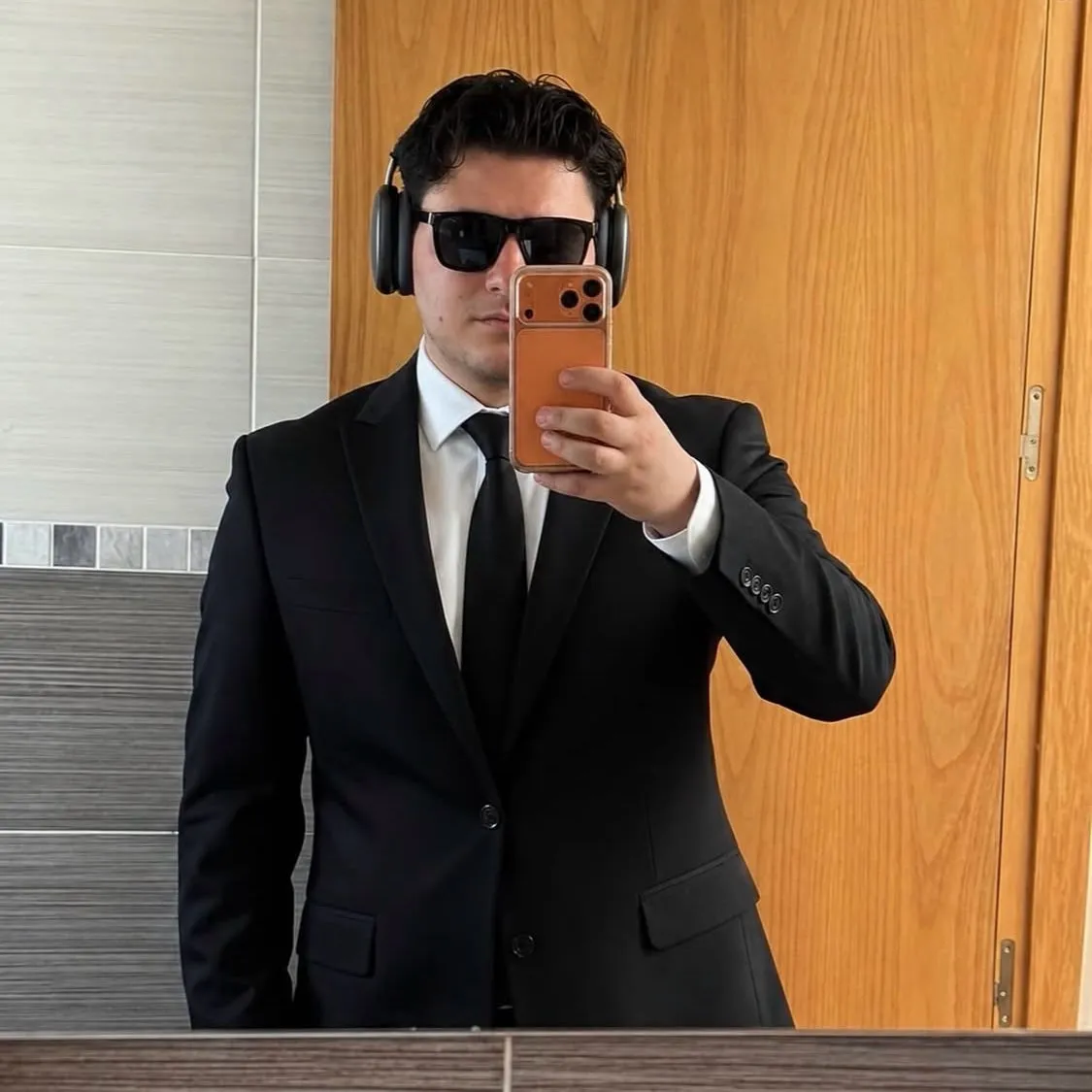 Aaron Alba con traje negro, gafas de sol y auriculares, haciéndose una foto en un espejo
