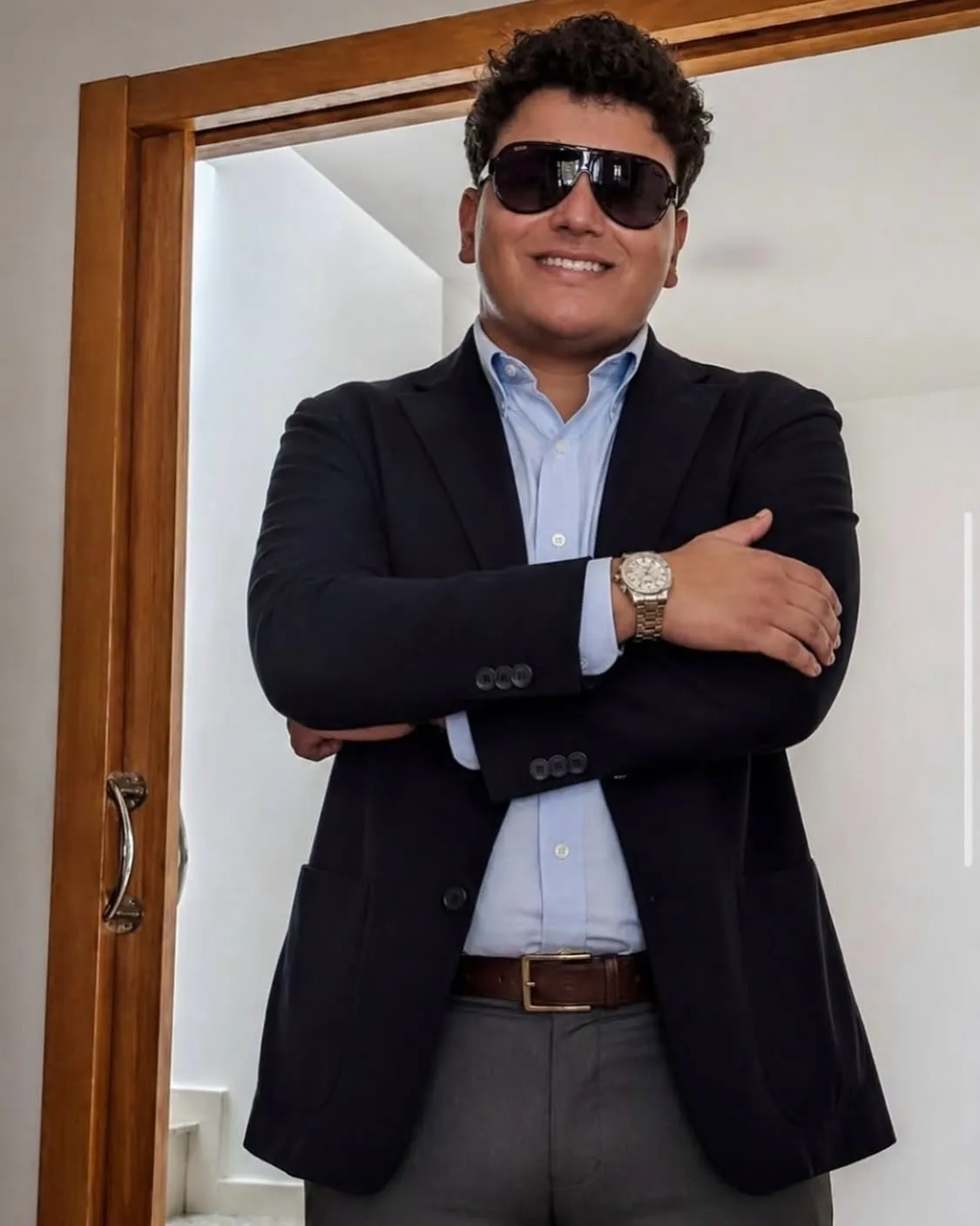 Aaron Alba de pie, sonriendo, con gafas de sol y los brazos cruzados junto a una puerta de madera